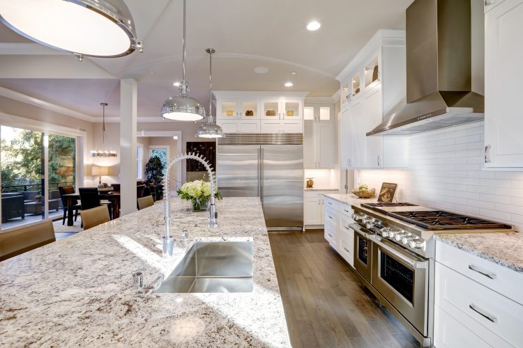 White kitchen design features large bar style kitchen island with granite countertop illuminated by modern pendant lights. Stainless steel appliances framed by white shaker cabinets . Northwest, USA